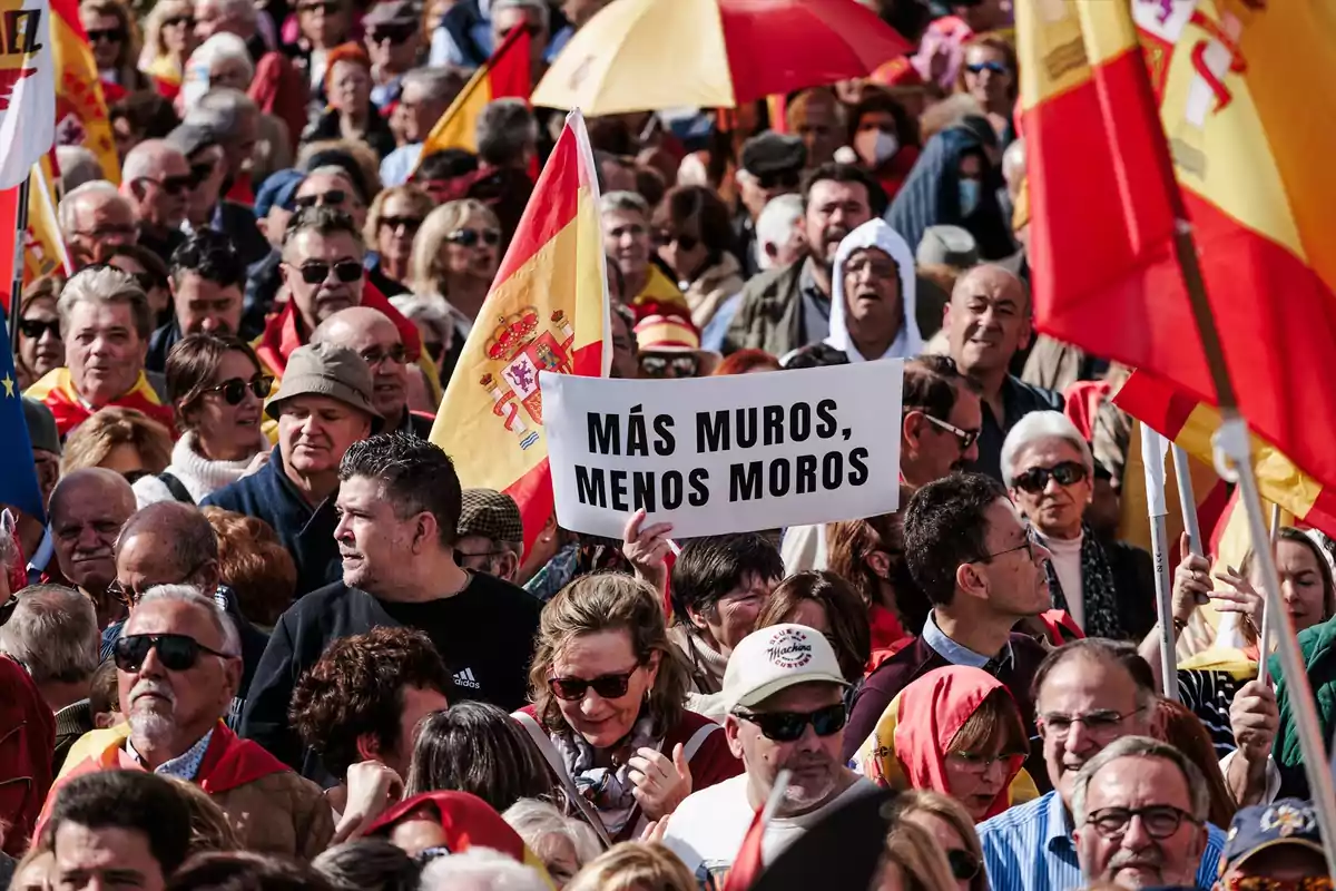 Un grupo de personas en una manifestación con banderas de España y un cartel con un mensaje controvertido.