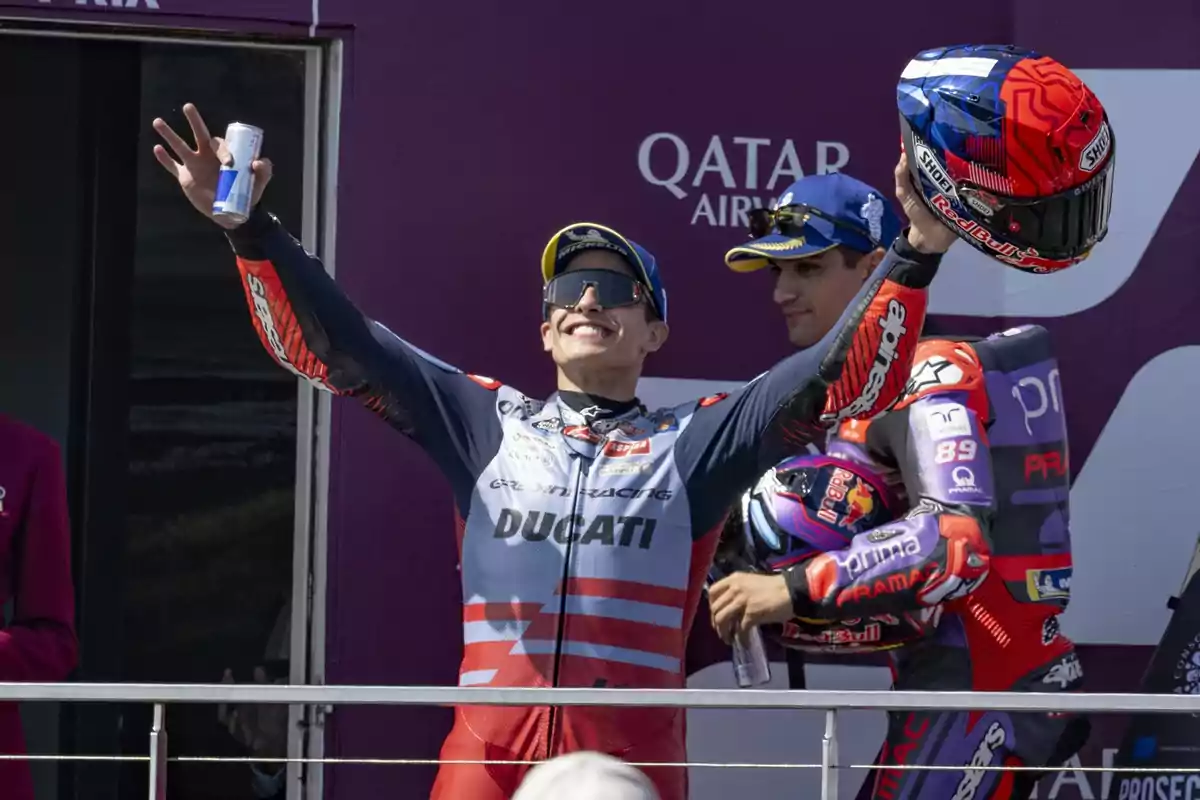 Dos pilotos de motociclismo celebran en el podio, uno de ellos levanta un casco y una bebida energética mientras sonríe.