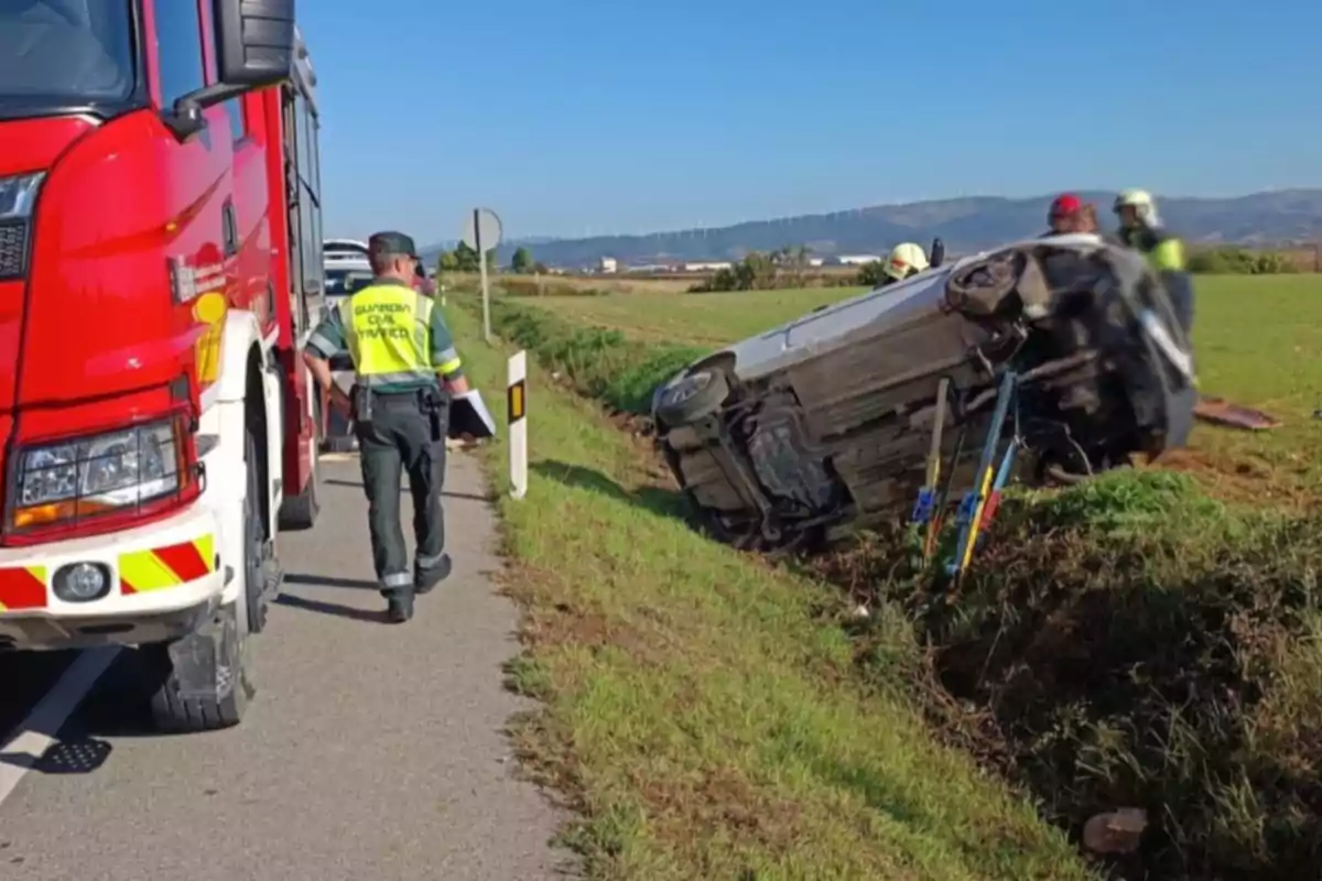 Un coche volcado en una zanja al lado de una carretera con un camión de bomberos y un agente de tráfico en la escena. Un coche volcado en una zanja al lado de una carretera con un camión de bomberos y un agente de tráfico en la escena.