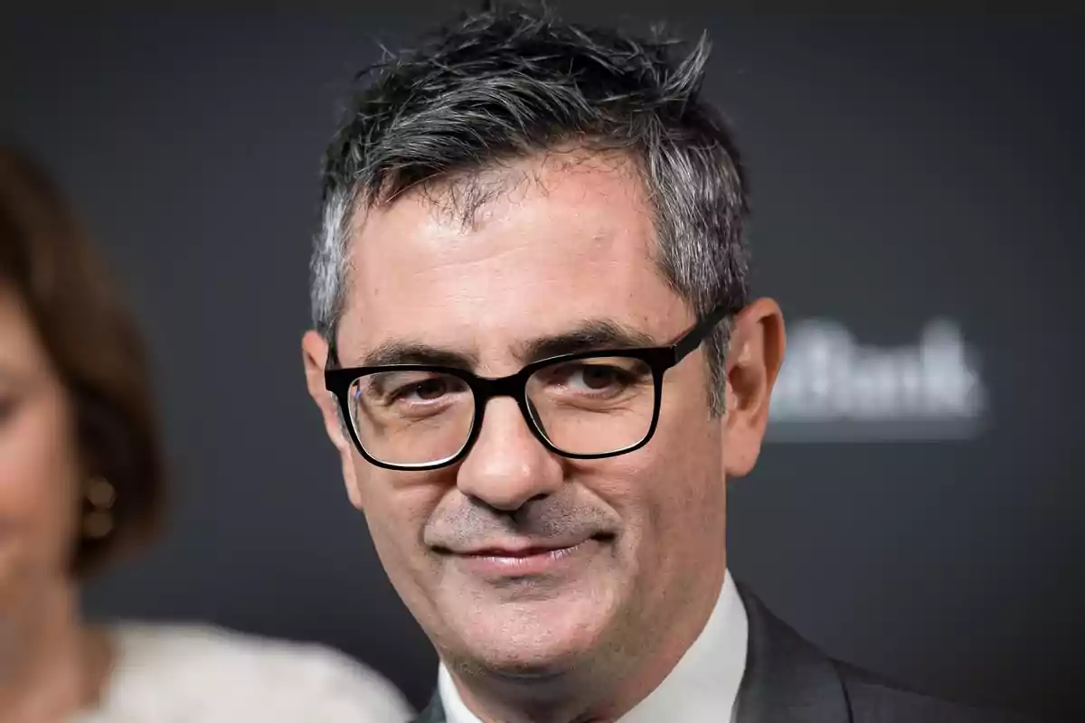 Un hombre con gafas y cabello canoso sonríe ligeramente mientras viste un traje formal.