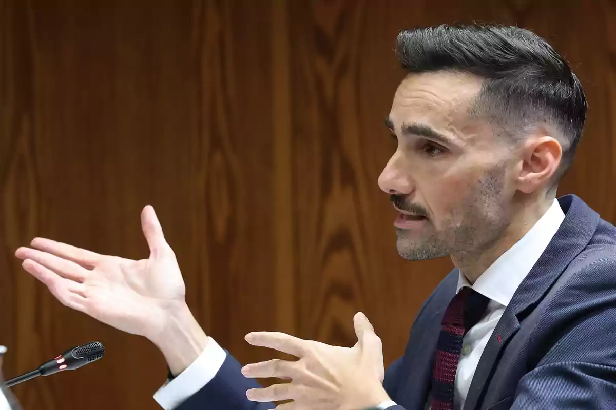 Un hombre de traje gesticula mientras habla en un entorno formal con un micrófono frente a él.