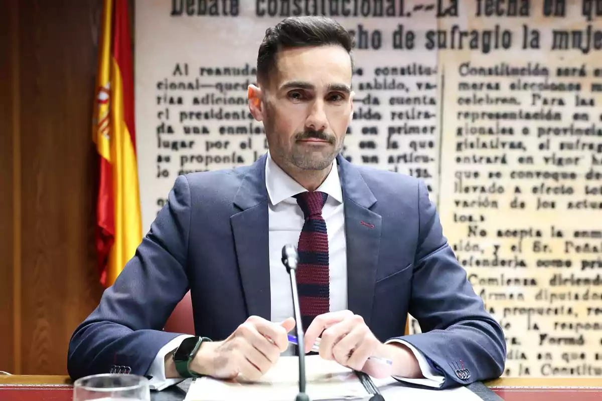 Hombre con traje y corbata sentado en una mesa frente a un micrófono con fondo de texto y una bandera de España Hombre con traje y corbata sentado en una mesa frente a un micrófono con fondo de texto y una bandera de España