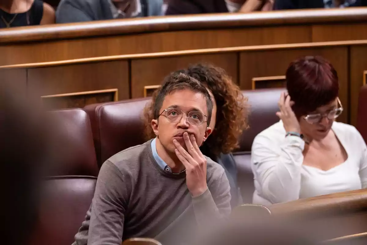 Una persona con gafas y suéter gris está sentada en un asiento de cuero en un entorno formal, con la mano en el rostro y expresión pensativa.