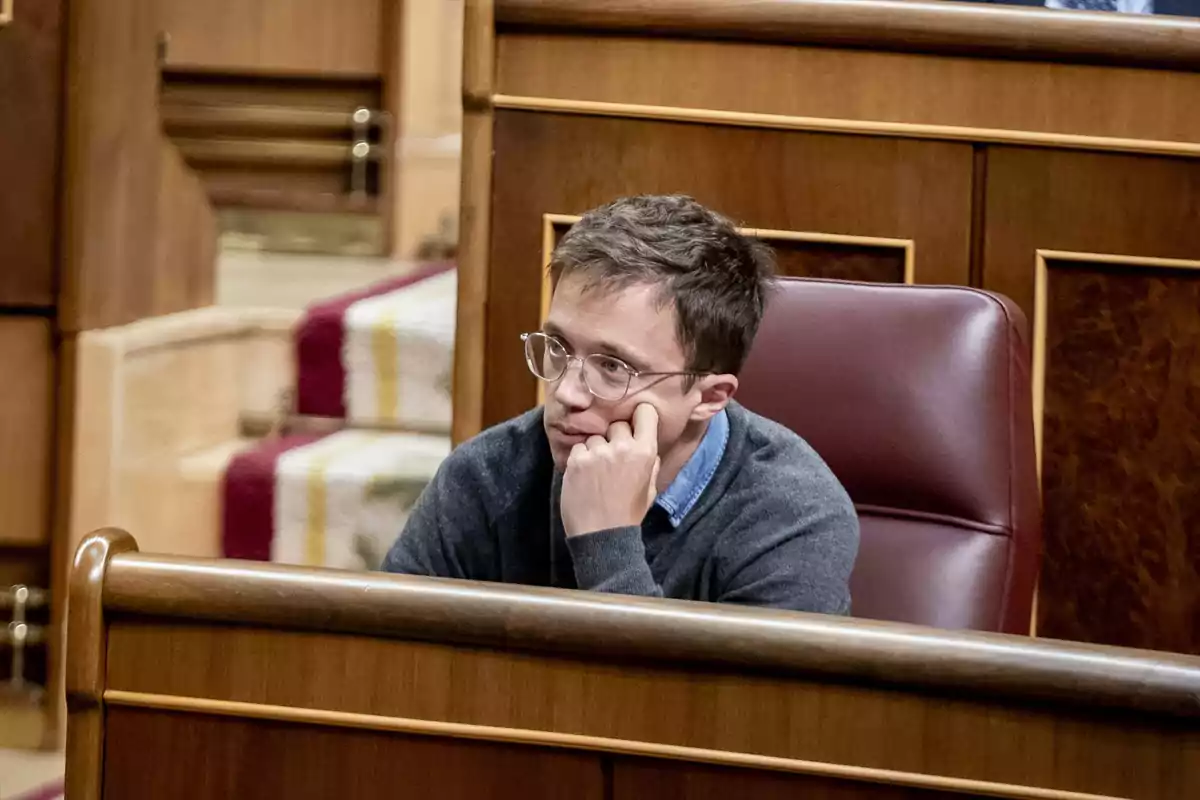 Una persona con gafas y suéter gris está sentada en un escaño de madera en un entorno formal.