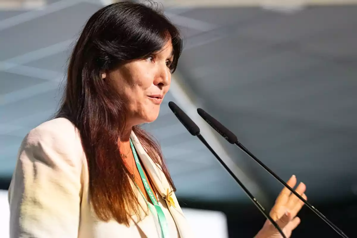 Mujer de cabello largo y oscuro hablando frente a dos micrófonos en un evento profesional