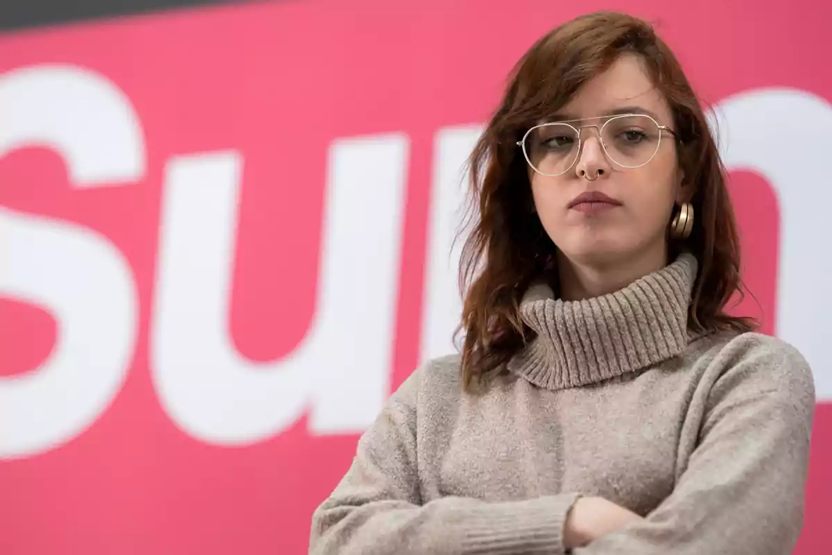 Una persona con gafas y suéter de cuello alto frente a un fondo rosa.