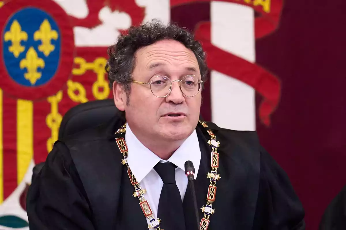 Un hombre con gafas y toga negra hablando frente a un micrófono con un fondo de escudos coloridos.