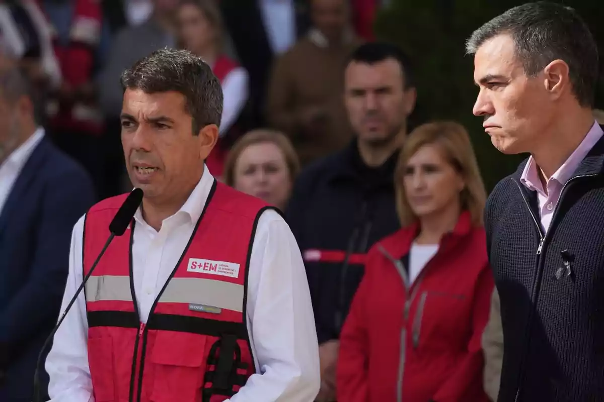 Un hombre con chaleco rojo habla frente a un micrófono mientras otras personas están de pie detrás de él.