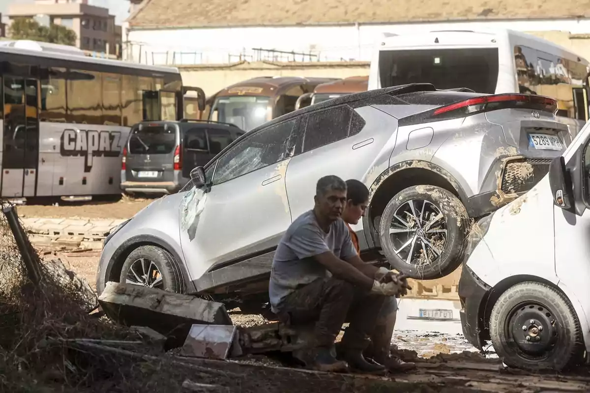 Un coche plateado está montado sobre otro vehículo en un área con varios autobuses estacionados y dos personas están sentadas en el suelo cerca de los vehículos.