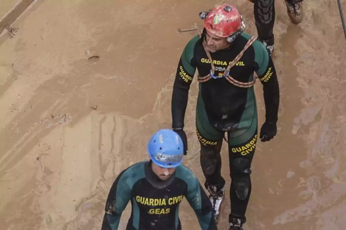 Dos miembros de la Guardia Civil con trajes de neopreno y cascos caminan por un terreno embarrado.