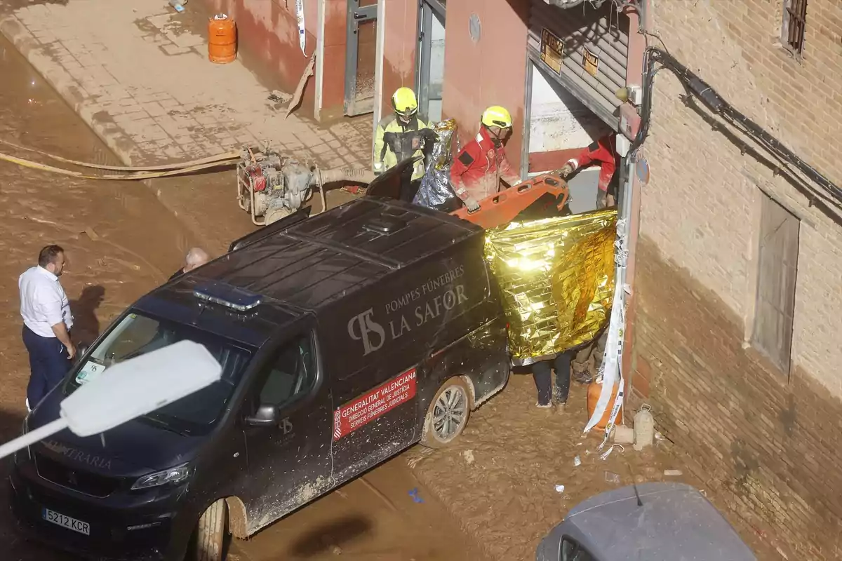 Un grupo de bomberos con cascos amarillos y rojos trabaja en una zona inundada junto a un vehículo de servicios funerarios mientras una persona observa la escena.