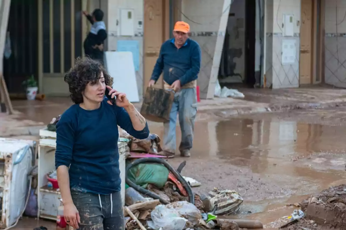 Una persona hablando por teléfono en una zona afectada por inundaciones con escombros y agua en el suelo mientras otra persona lleva un objeto pesado.