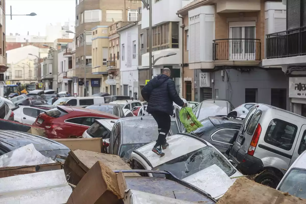 Un hombre camina sobre autos dañados en una calle llena de escombros y vehículos amontonados tras una inundación.