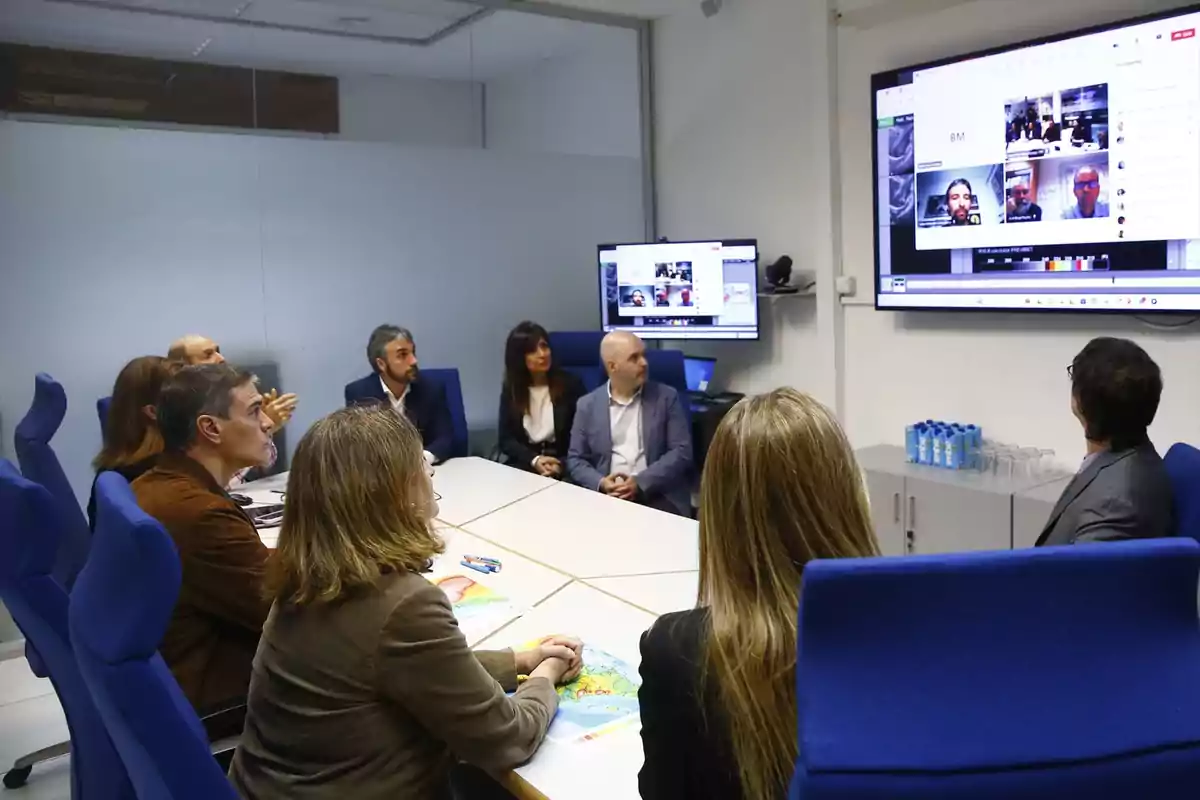 Un grupo de personas en una sala de conferencias participando en una videollamada, con pantallas mostrando a otros participantes.