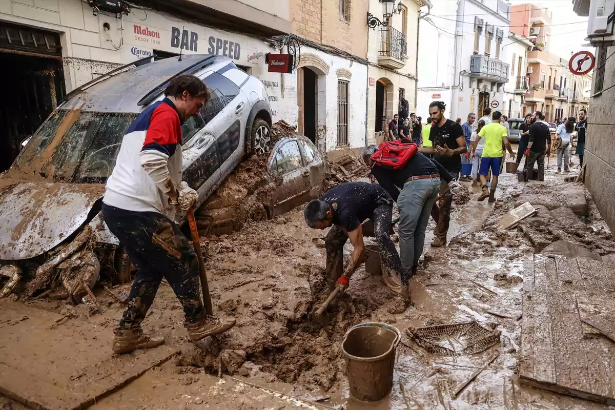 Personas trabajando para limpiar una calle llena de barro y escombros tras una inundación, con un coche dañado en el fondo.