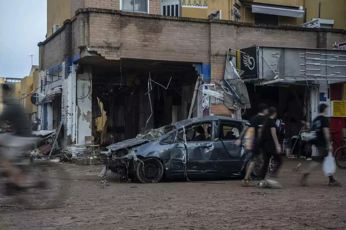 Un coche dañado frente a un edificio con fachada destruida y personas caminando por una calle cubierta de barro.