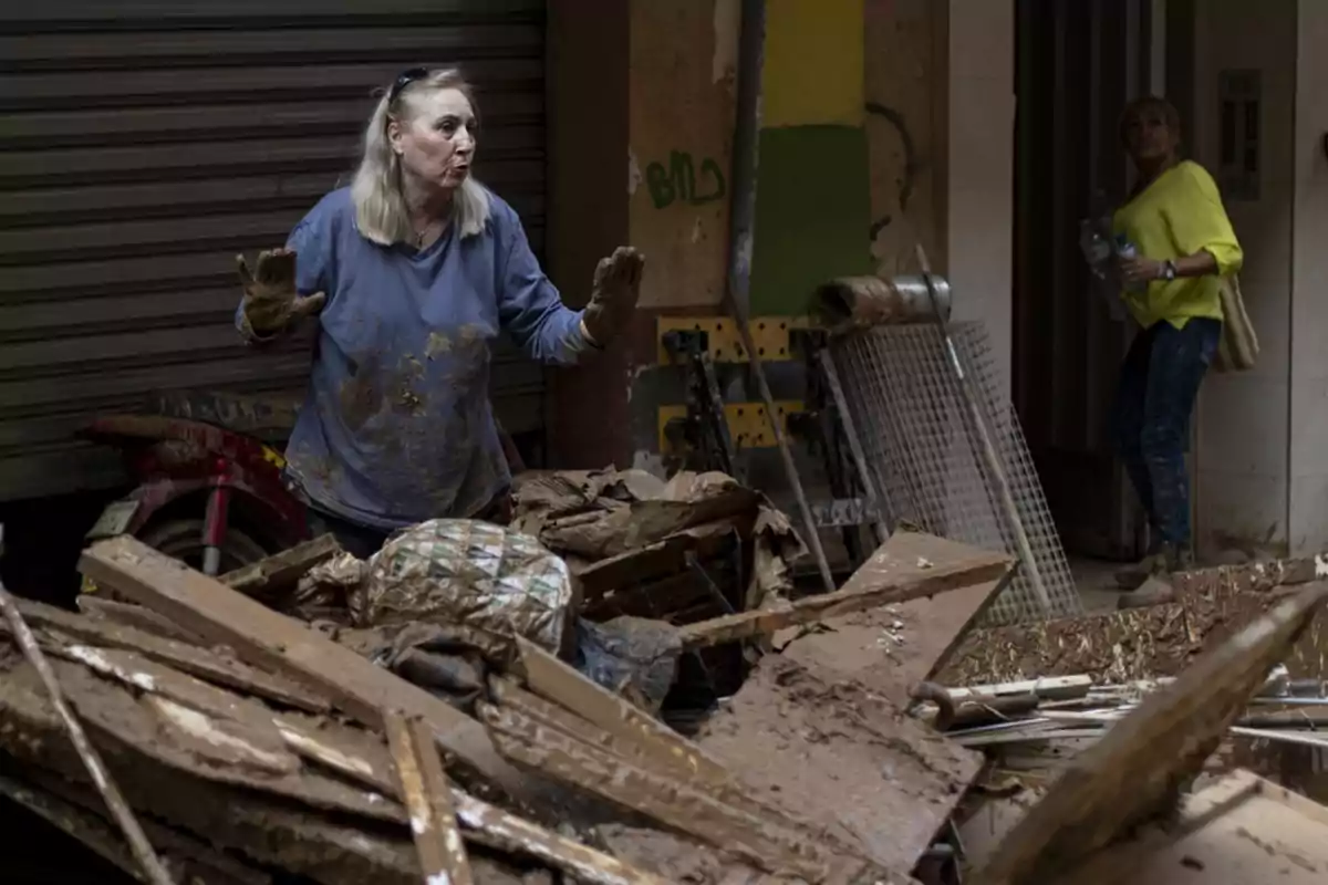 Una mujer con ropa embarrada gesticula mientras está rodeada de escombros y barro en un entorno urbano.