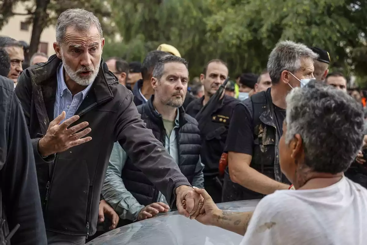 Un hombre de cabello canoso y barba estrecha la mano de una persona en un entorno al aire libre rodeado de varias personas, algunas de las cuales llevan mascarillas.