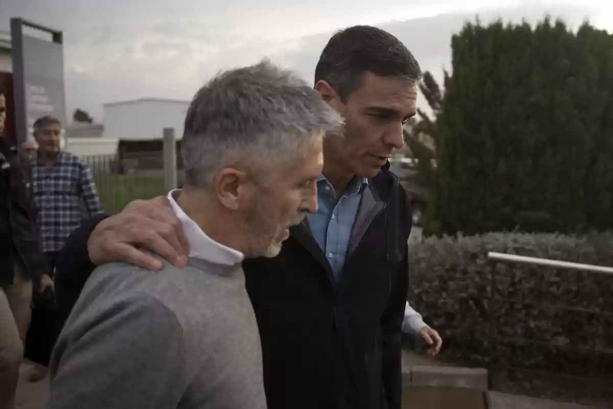 Dos hombres caminando juntos mientras uno de ellos tiene el brazo sobre el hombro del otro en un entorno al aire libre con vegetación y un edificio al fondo.