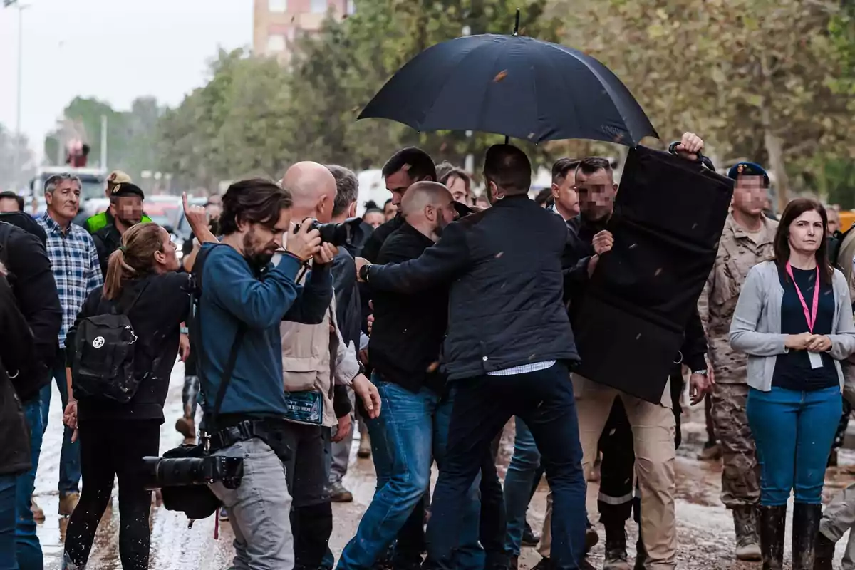 Un grupo de personas, incluyendo fotógrafos y personal de seguridad, se encuentra en una calle mientras uno de ellos sostiene un paraguas negro.