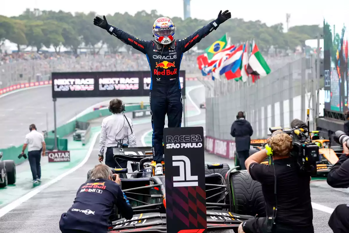 Piloto de Fórmula 1 celebrando sobre su auto en el podio del Gran Premio de São Paulo con fotógrafos capturando el momento y banderas de varios países al fondo.