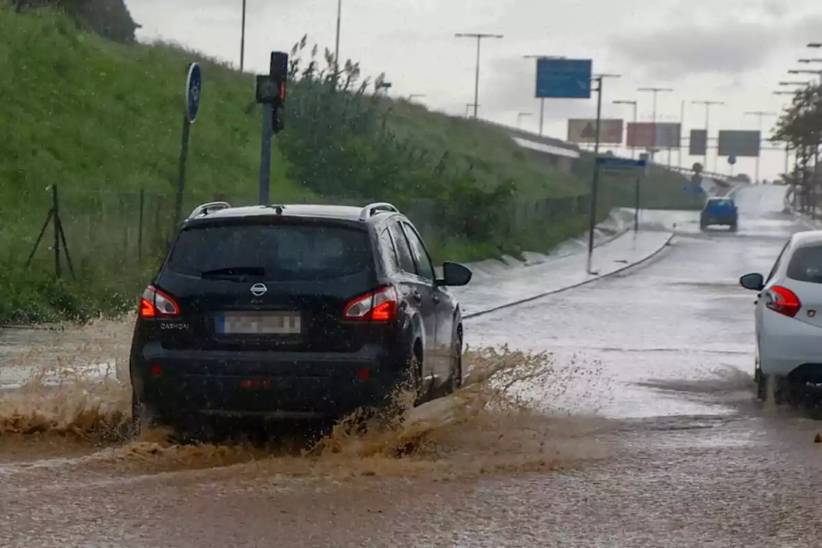 Un coche negro circula por una carretera inundada mientras el agua salpica a su alrededor en un día lluvioso.