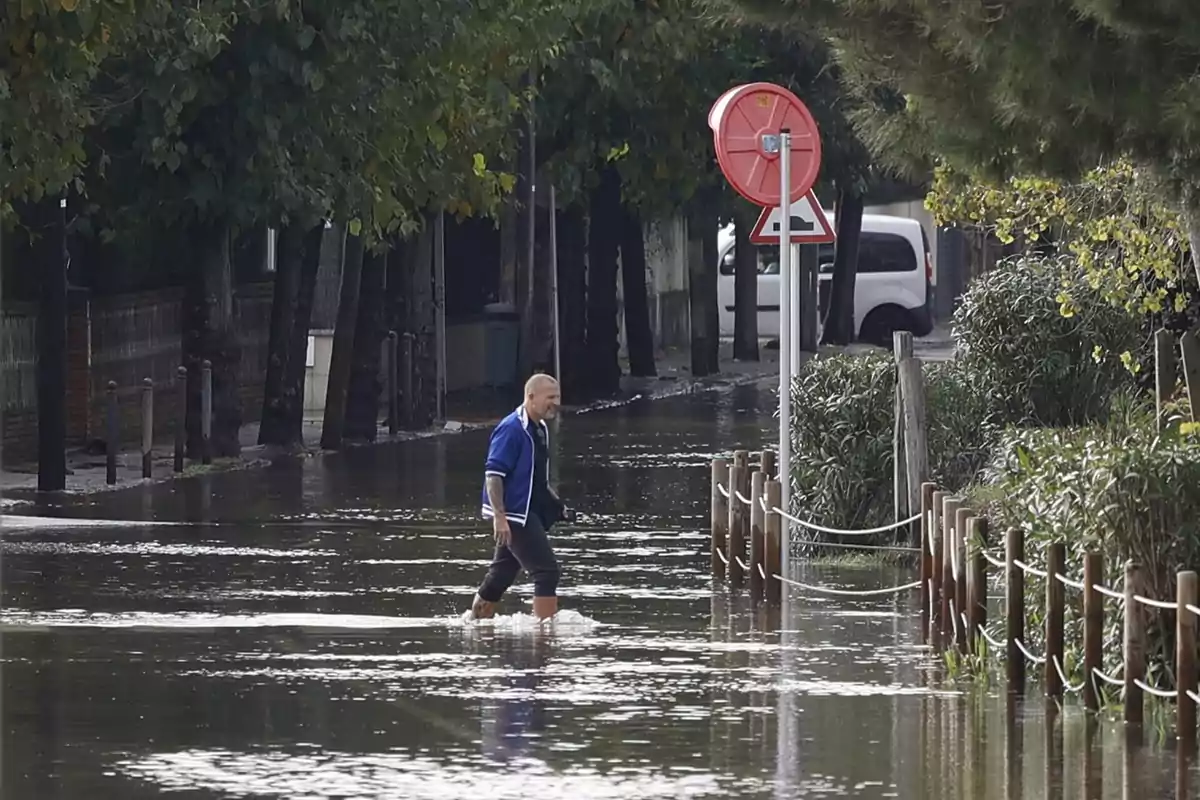 Un hombre camina por una calle inundada con agua hasta las rodillas, rodeado de árboles y un vehículo estacionado al fondo.