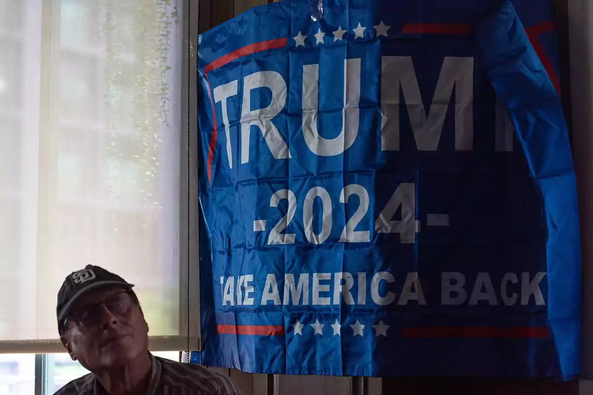 Un hombre con gorra y gafas está sentado junto a una bandera azul con texto político.