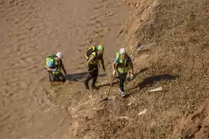 Tres personas con equipo de protección y cascos caminan por un terreno fangoso junto a un río.