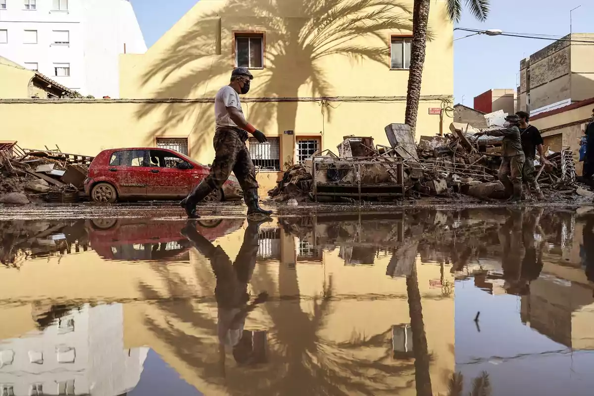 Un hombre camina por una calle inundada con escombros y un coche rojo al fondo reflejados en el agua.