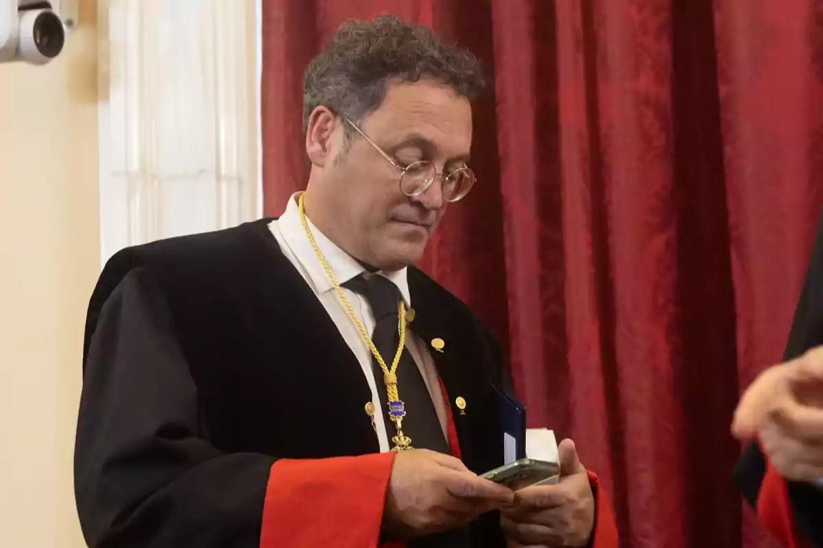 A man in an academic gown and a gold medal holds an object in his hands in front of a red curtain.