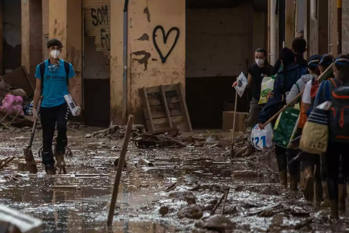 Personas con mascarillas caminan por una calle inundada y llena de barro, llevando herramientas y bolsas, mientras un edificio con un corazón pintado en la pared se ve al fondo.