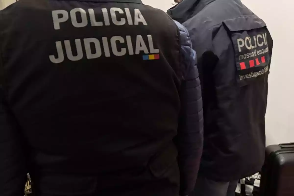 Dos agentes de policía vestidos con chalecos identificativos de la policía judicial y de investigación de los Mossos d'Esquadra están de espaldas en una habitación. Dos agentes de policía vestidos con chalecos identificativos de la policía judicial y de investigación de los Mossos d'Esquadra están de espaldas en una habitación.