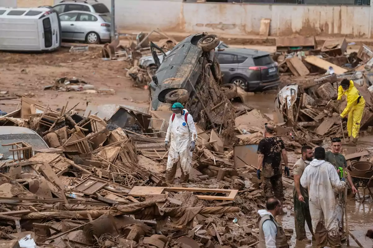 Personas con trajes protectores caminan entre escombros y vehículos volcados tras un desastre natural.