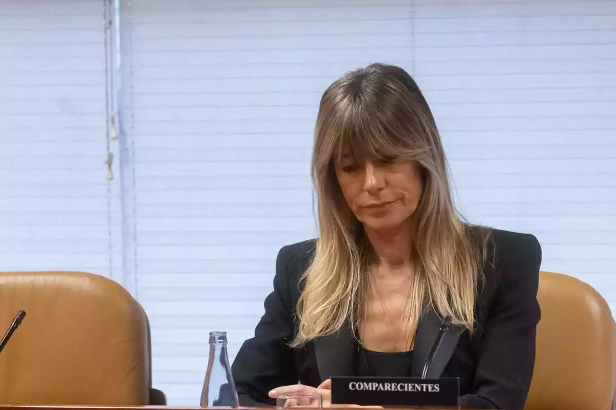 A woman with blonde hair and bangs sits in a conference room, dressed in a dark suit, in front of a sign that says 