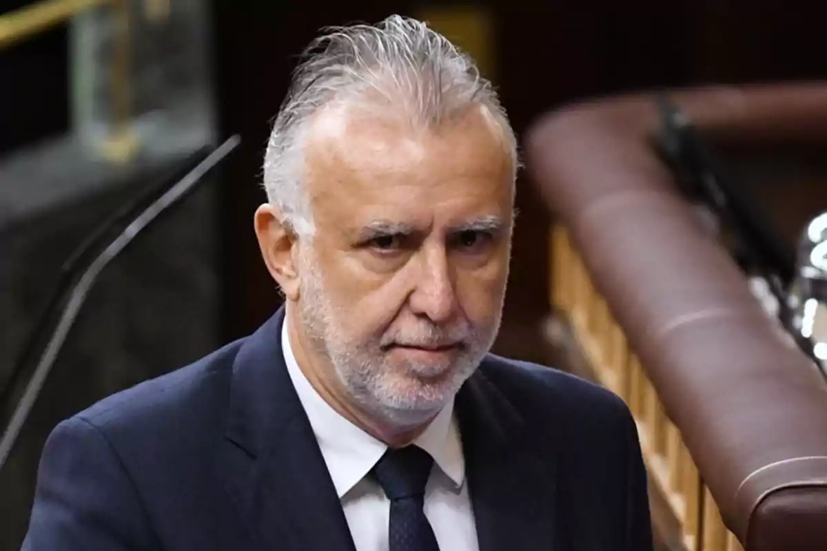 Un hombre de cabello canoso y barba vestido con traje oscuro y corbata en un entorno formal.