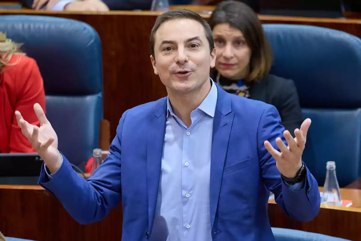 Un hombre con traje azul gesticula mientras habla en un entorno formal con personas sentadas detrás de él.