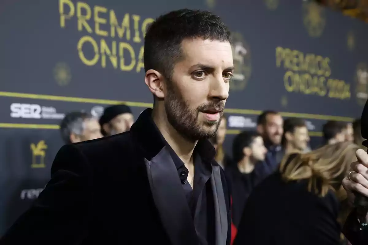 Hombre con barba y cabello corto vestido con traje negro en la alfombra de los Premios Ondas con fondo de personas y logotipos del evento. Hombre con barba y cabello corto vestido con traje negro en la alfombra de los Premios Ondas con fondo de personas y logotipos del evento.