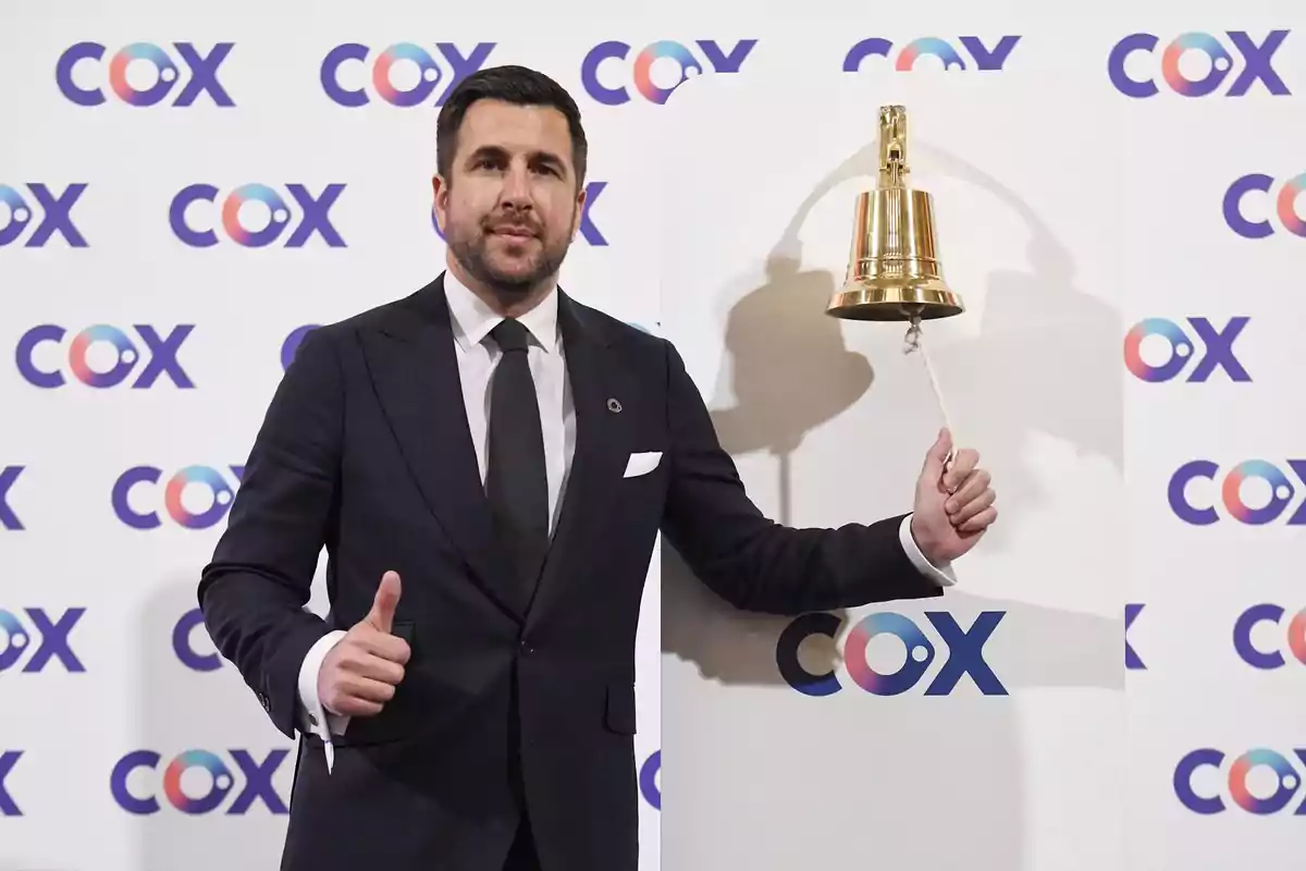 Un hombre en traje oscuro toca una campana dorada frente a un fondo con el logo de COX.