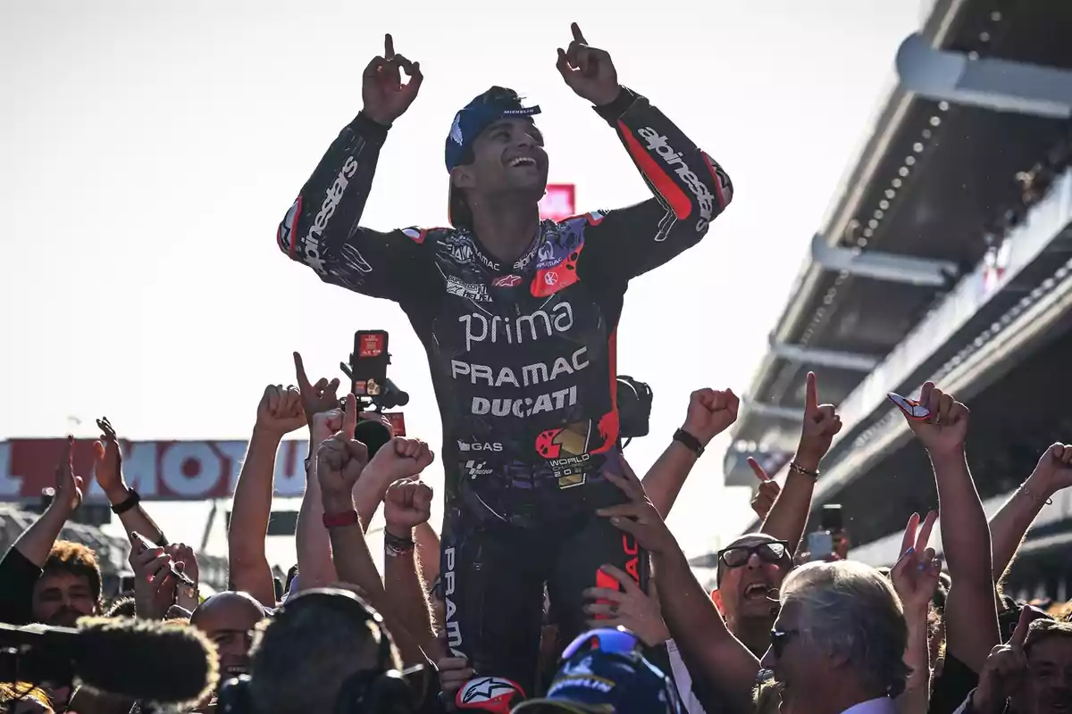 Un piloto de motociclismo celebra levantado por una multitud de personas que lo vitorean y aplauden en un circuito de carreras.