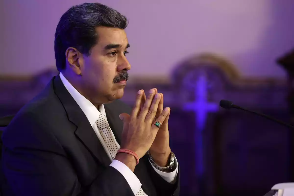 Un hombre de traje oscuro y corbata clara está sentado con las manos juntas frente a un micrófono en un entorno iluminado con luz púrpura.