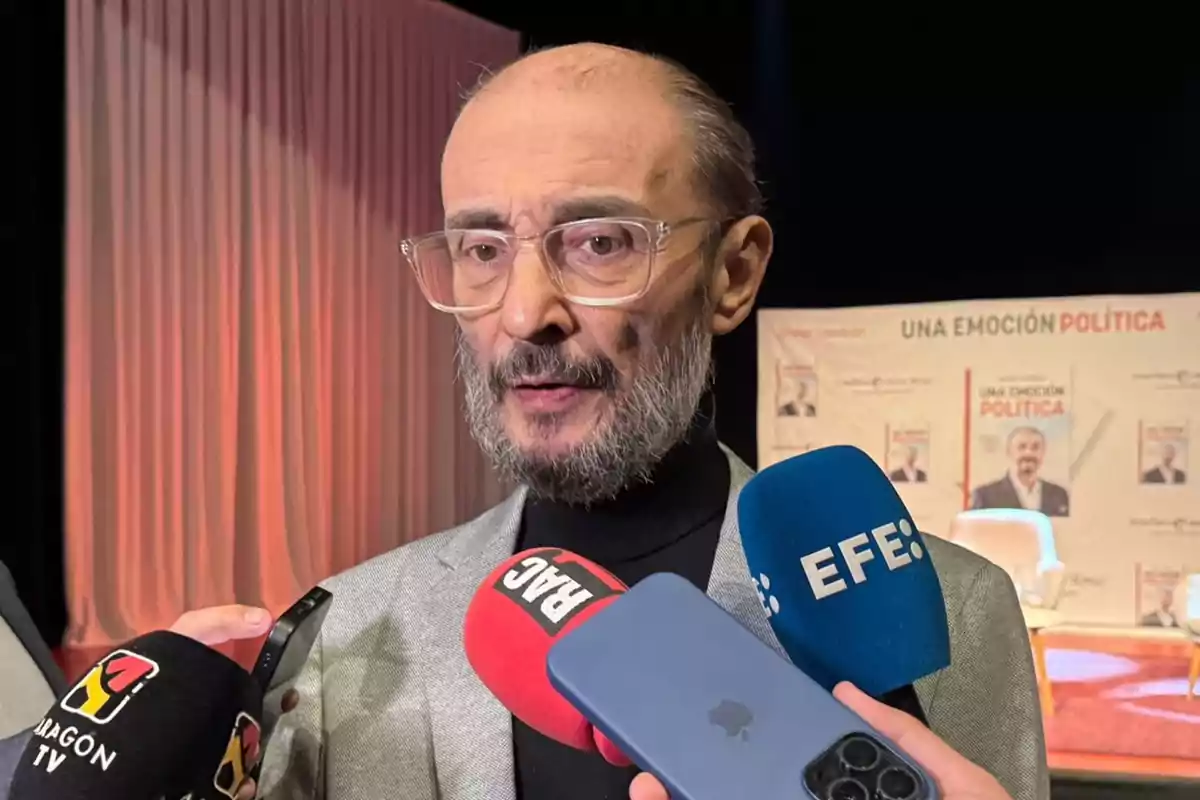 Hombre de barba y gafas transparentes hablando con la prensa mientras sostiene varios micrófonos frente a él en un evento de presentación de un libro titulado Una emoción política Hombre de barba y gafas transparentes hablando con la prensa mientras sostiene varios micrófonos frente a él en un evento de presentación de un libro titulado Una emoción política