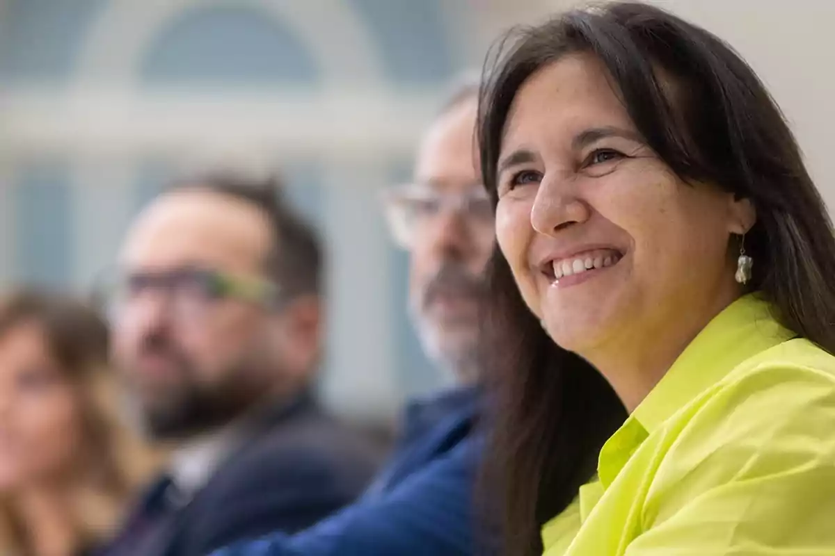 Una mujer sonriente con una camisa amarilla está sentada en un evento, con otras personas desenfocadas en el fondo.