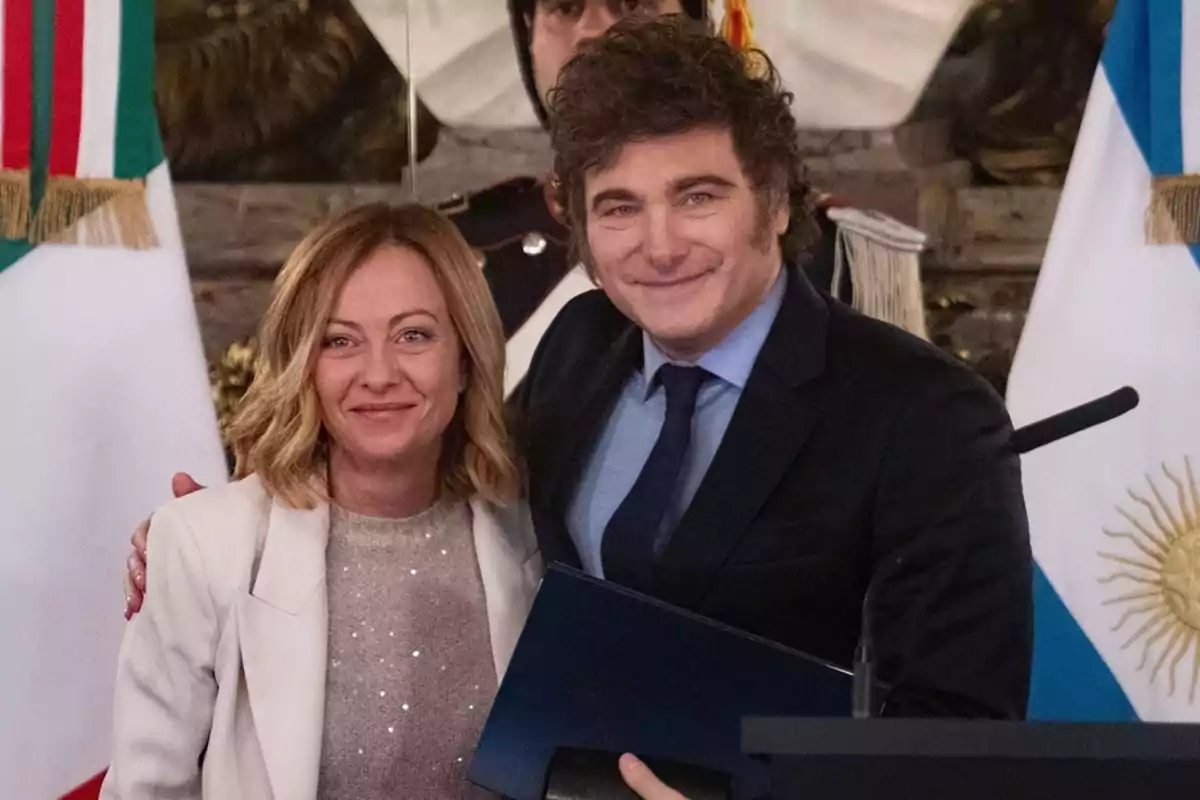 Dos personas posan juntas sonriendo frente a banderas de Italia y Argentina.