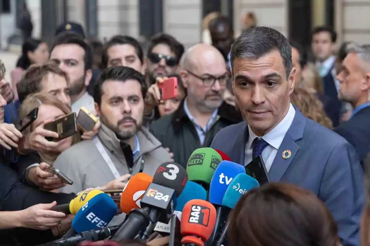 Un hombre en traje rodeado de periodistas y micrófonos de diferentes medios de comunicación.