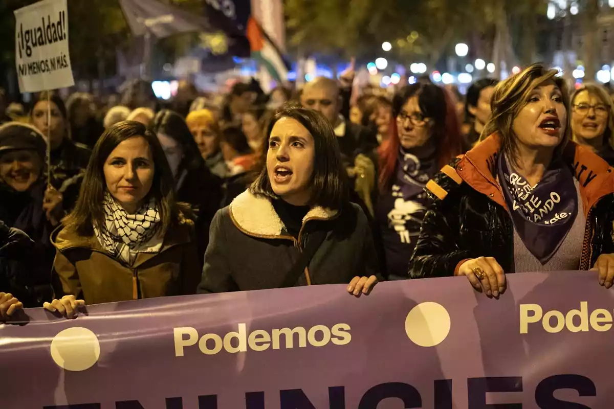 Un grupo de personas participa en una manifestación nocturna sosteniendo una pancarta morada con el logo de un partido político y un cartel que pide igualdad.