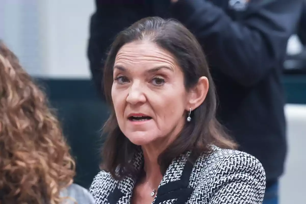 Mujer de cabello oscuro y chaqueta de tweed hablando en un entorno interior.