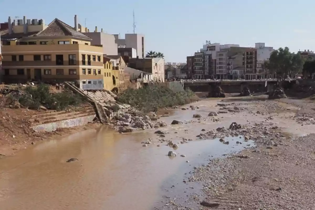 Vista de un río con poca agua y piedras en el cauce rodeado de edificios y vegetación.