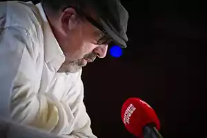 Hombre con gafas y gorra hablando frente a un micrófono rojo.