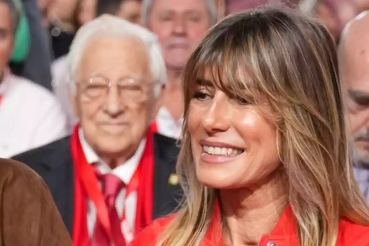 Una mujer sonriente con cabello rubio y flequillo en un evento con personas de fondo.